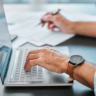 Eine Hand, die auf der Tastatur eines Laptops liegt, im Hintergrund eine andere Hand, die etwas notiert 